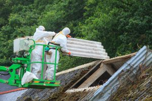 Two workers in protective suits removing asbestos roofing sheets from a building using a mobile platform.